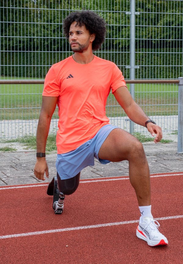 Uomo con una gamba protesica che indossa una maglietta arancione e pantaloncini blu mentre si allunga su una pista all'aperto vicino a una recinzione di metallo.