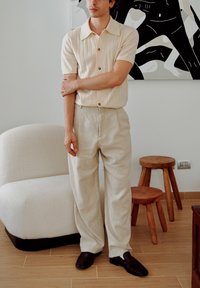 Camisa de manga curta bege com riscas e botões castanhos, combinada com calças de linho bege claro. Sapatos escuros, de design simplista, contra um fundo minimalista.