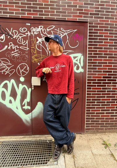 Camisa corta de manga larga roja con logo, gorra azul, vaqueros holgados azul oscuro y zapatos negros, de pie contra una pared cubierta de grafitis.