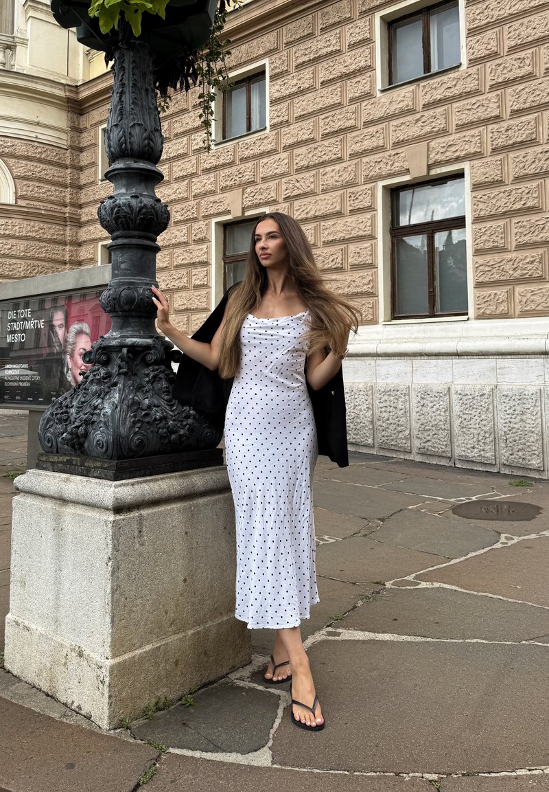 Femme aux longs cheveux portant une robe blanche à pois et une veste noire, se tient sur le trottoir de la ville en tenant un lampadaire orné à côté d'un bâtiment historique.
