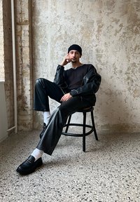Melnā ādas jaka un bikses, ribota melna džemperis, melni loafers ar ķēdes detaļu, baltas zeķes, pozējot sēžot uz koka krēsla.