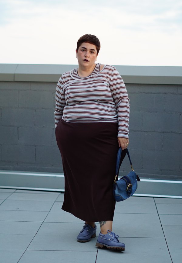 Person with short dark hair wearing a striped long-sleeve top, dark skirt, blue shoes, holding a blue handbag, standing outdoors on tiled floor.