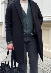 Fekete kabát zöld kötött kardigán felett, amelynek kék gombjai vannak, szürke farmerrel viselve, és egy fekete táskát tartva. Láthatóak a texturált anyag részletei.