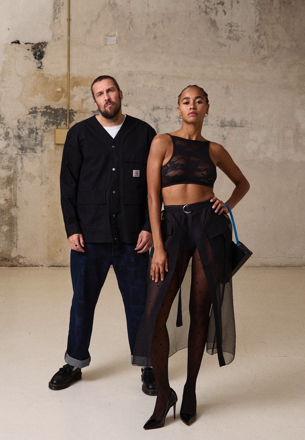 Top cropped de renda preta e saia fluida transparente com fendas na coxa; homem com camisa preta de botões, calças azuis com padrão e sapatos escuros. Acessórios de bolsa minimalista.