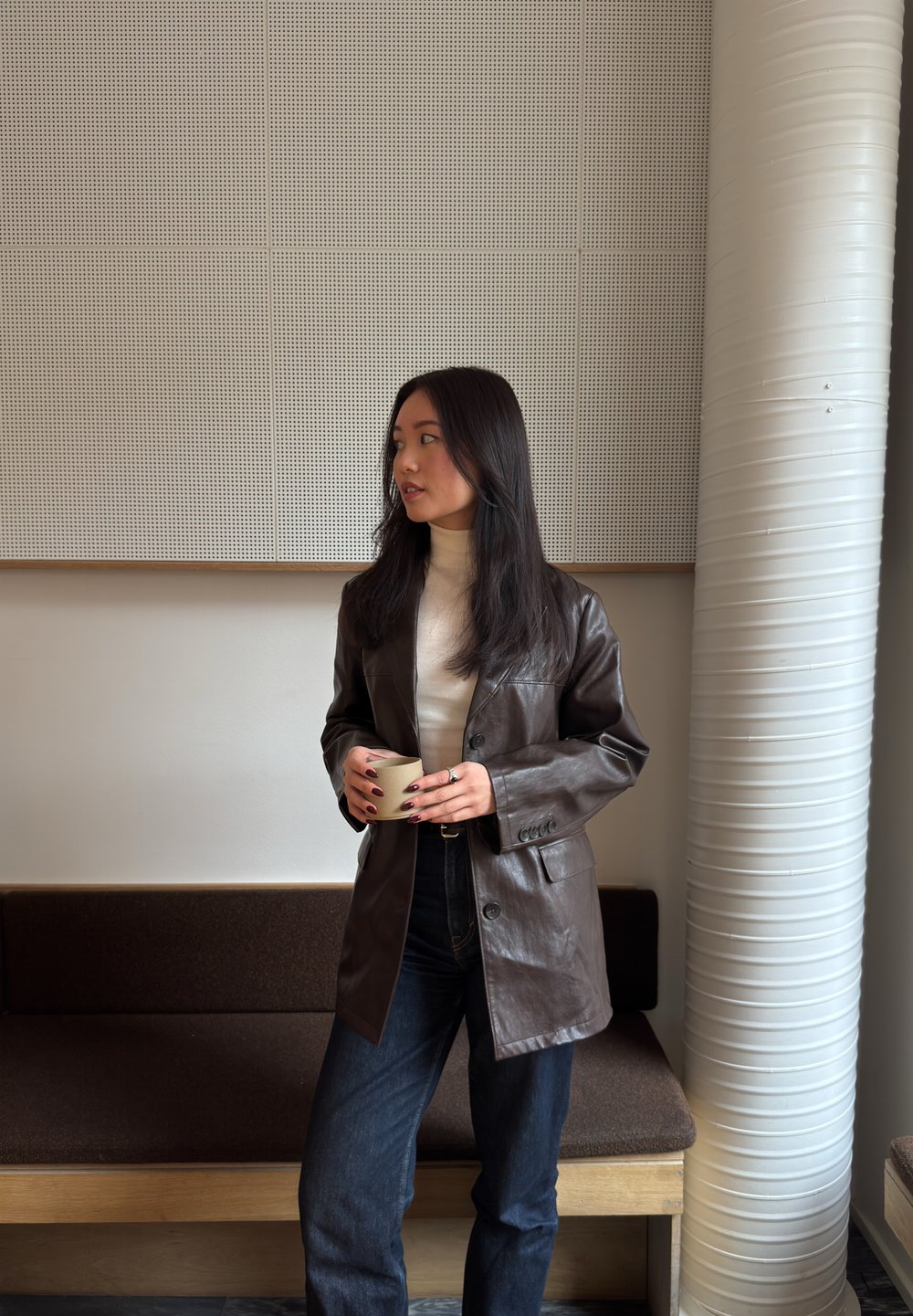 Jeune femme portant une veste en cuir marron et un col roulé blanc, tenant une tasse, debout à l'intérieur près d'un banc et d'une grande colonne blanche.