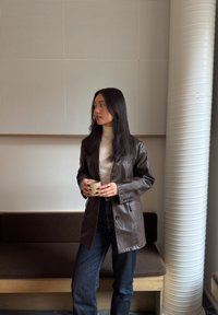 Jeune femme portant une veste en cuir marron et un col roulé blanc, tenant une tasse, debout à l'intérieur près d'un banc et d'une grande colonne blanche.