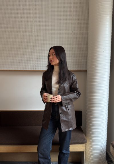 Joven mujer con chaqueta de cuero marrón y suéter blanco de cuello alto sosteniendo una taza, de pie en el interior junto a un banco y una gran columna blanca.