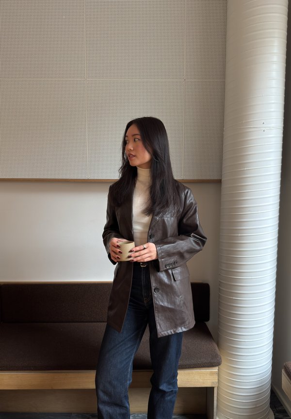 Tânără în geacă maro din piele și pulover cu guler înalt alb, ținând o cană, stând înăuntru lângă o bancă și o coloană mare albă.