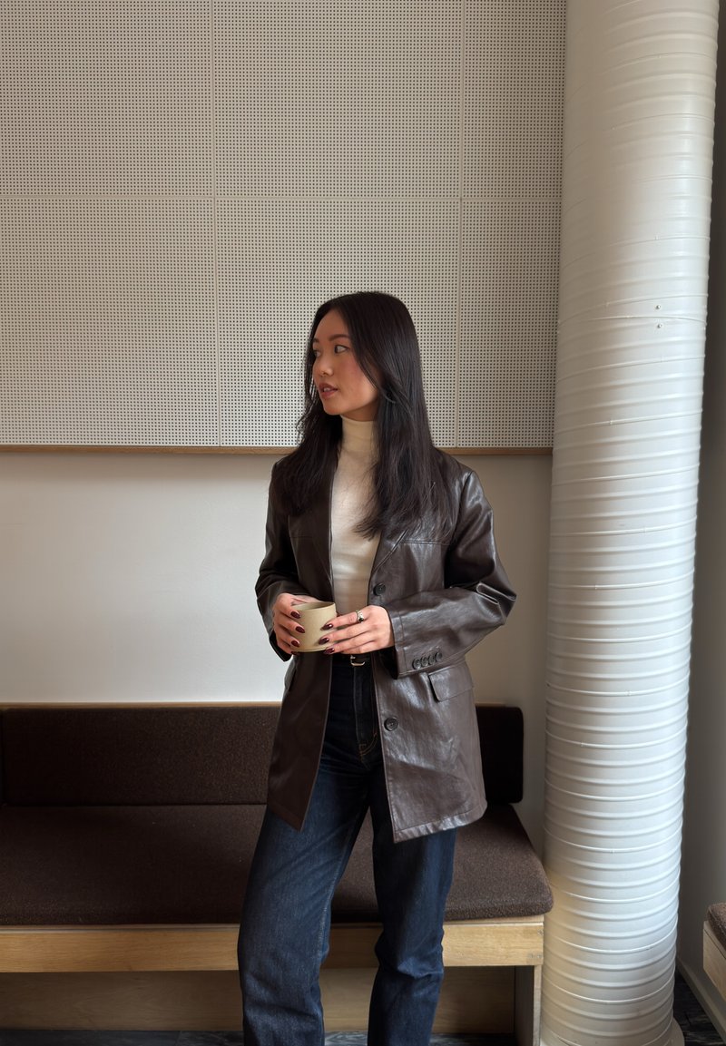 Jeune femme portant une veste en cuir marron et un col roulé blanc, tenant une tasse, debout à l'intérieur près d'un banc et d'une grande colonne blanche.