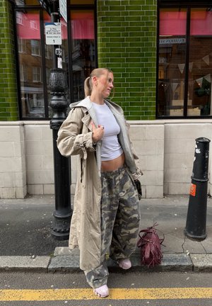 Joven mujer con abrigo beige, top blanco recortado y pantalones de camuflaje, de pie en una acera de la ciudad junto a un edificio con azulejos verdes y una bolsa roja.