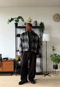 Veste courte à carreaux noir et blanc avec col, portée sur une chemise foncée, associée à un pantalon noir ample et des chaussures noires. Cadre intérieur avec des plantes.