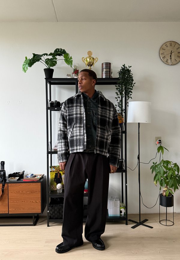 Veste courte à carreaux noir et blanc avec col portée sur une chemise foncée, associée à un pantalon noir ample et des chaussures noires. Décor intérieur avec des plantes.