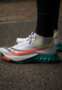 Scarpe sportive bianche con tomaia texturizzata, caratterizzate da vivaci dettagli arancioni e verdi e una suola seghettata color teal.