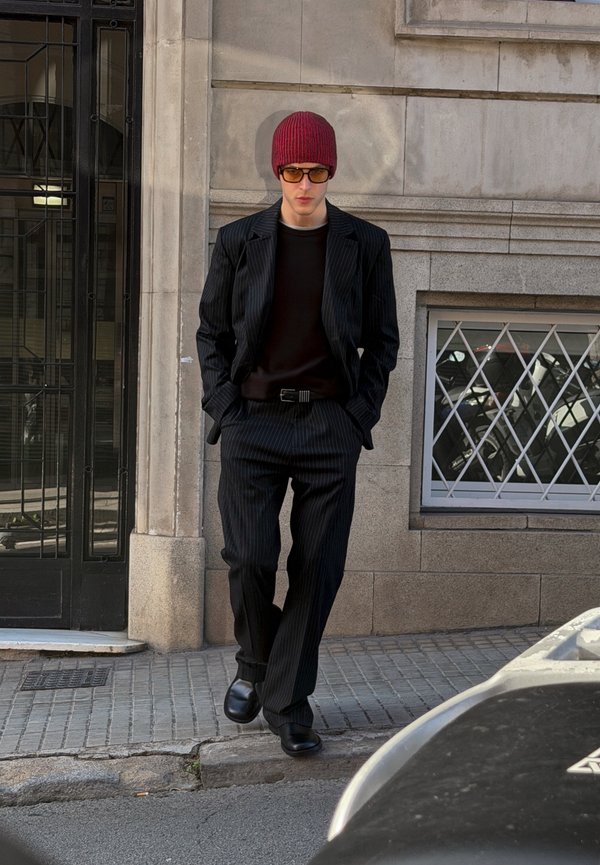 Bărbat purtând costum negru cu dungi, cămașă neagră, cagulă roșie și ochelari de soare, mergând pe trotuarul unui oraș lângă o clădire cu fereastră împrejmuită cu gratii.