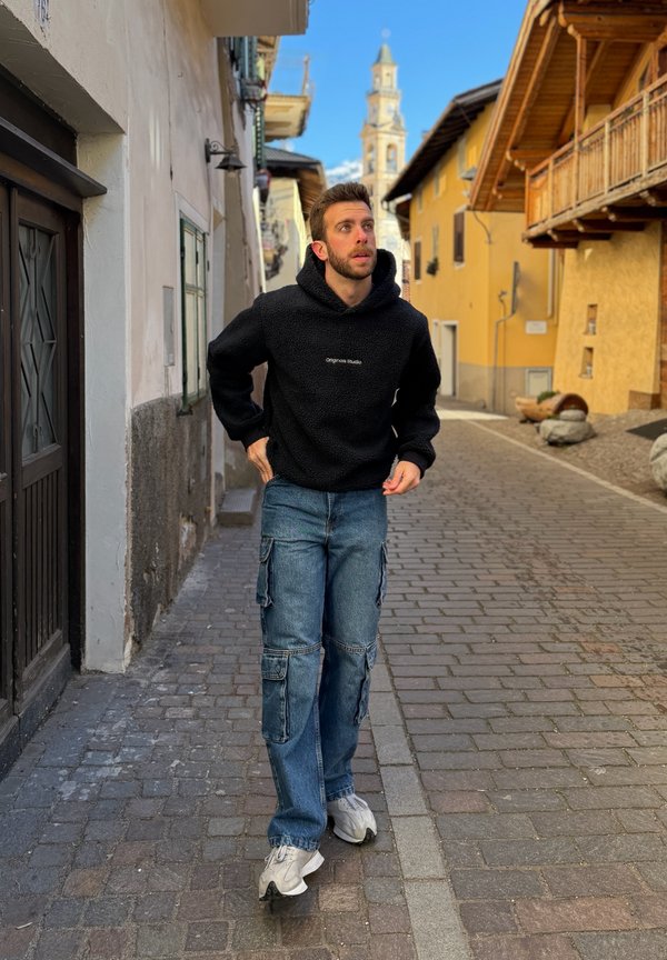 Giovane uomo con felpa nera con cappuccio e jeans che cammina su una strada lastricata con edifici gialli e una torre con orologio sullo sfondo.
