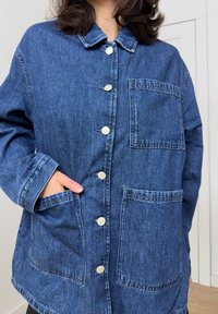 Džínová bunda tmavě modré barvy s límečkem, předním zapínáním na knoflíky, dvěma velkými nakládanými kapsami a volným střihem.