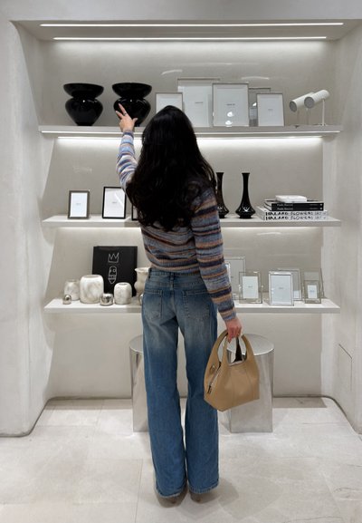 Mujer con suéter de rayas y vaqueros alcanza jarrones de cerámica negra en estantes blancos iluminados que exhiben marcos, libros y piezas decorativas.