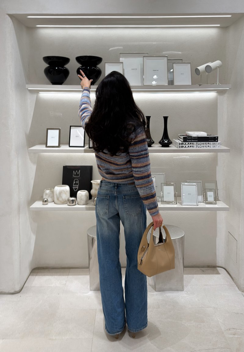 Femme en pull rayé et jeans attrape des vases en céramique noire sur des étagères blanches éclairées présentant des cadres, des livres et des objets décoratifs.