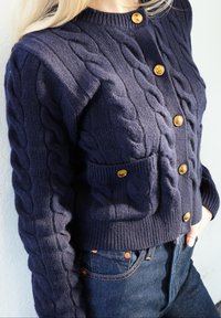 Tmavomodrý kardigán s vrkočovým vzorom a rebrovanou textúrou, ozdobený zlatými gombíkmi a vreckom vpredu. Nosený cez tmavé riflové nohavice.