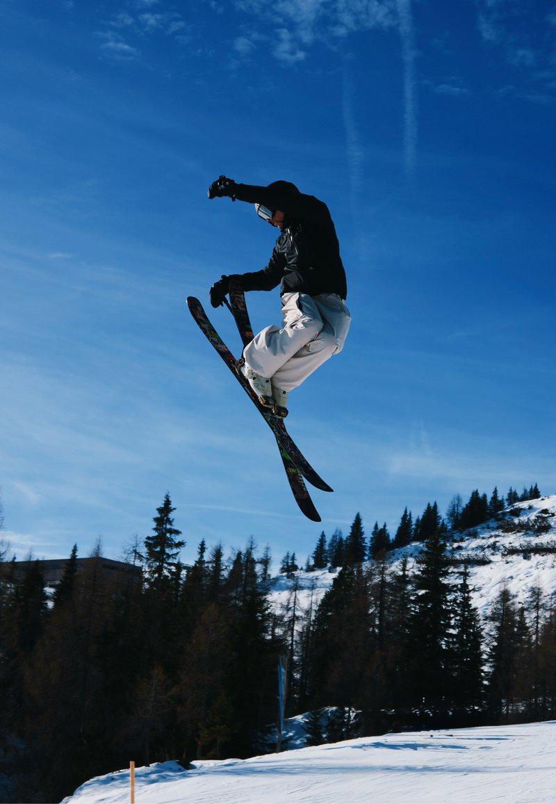 Un skieur en tenue noire et pantalon blanc effectue un saut avec des skis colorés sur fond de ciel bleu, avec des arbres enneigés en arrière-plan.