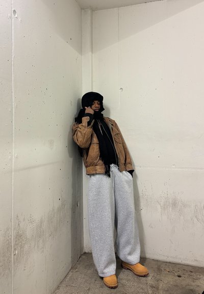 Persona vestida con una chaqueta marrón, bufanda negra y capucha, pantalones de chándal grises y botas beige, apoyada contra una pared de hormigón manchada en un rincón.