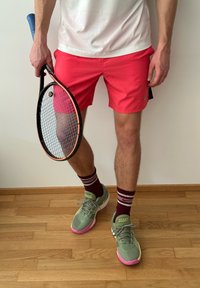 Conjunto de ténis masculino: calções cor-de-rosa brilhantes, camiseta branca, sapatilhas verdes com detalhes cor-de-rosa, meias bordeaux com riscas e segurando uma raquete de ténis.