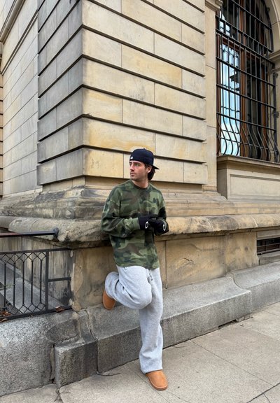 Camisa de manga larga de camuflaje verde, pantalones de chándal grises y botas marrones. Guantes negros con un acento circular. Apoyado contra una pared de piedra.