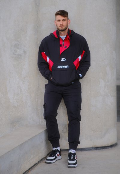 Jersey pulóver negro y rojo con logo, combinado con pantalones cargo negros y zapatillas blancas con acentos rojos. Textura suave y diseño deportivo.