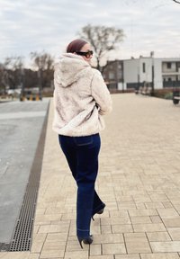 Beiger Kapuzenpullover aus Kunstpelz mit Kapuze, kombiniert mit dunkelblauen Schlagjeans und schwarzen High Heels, vor einem gepflasterten Außenbereich.