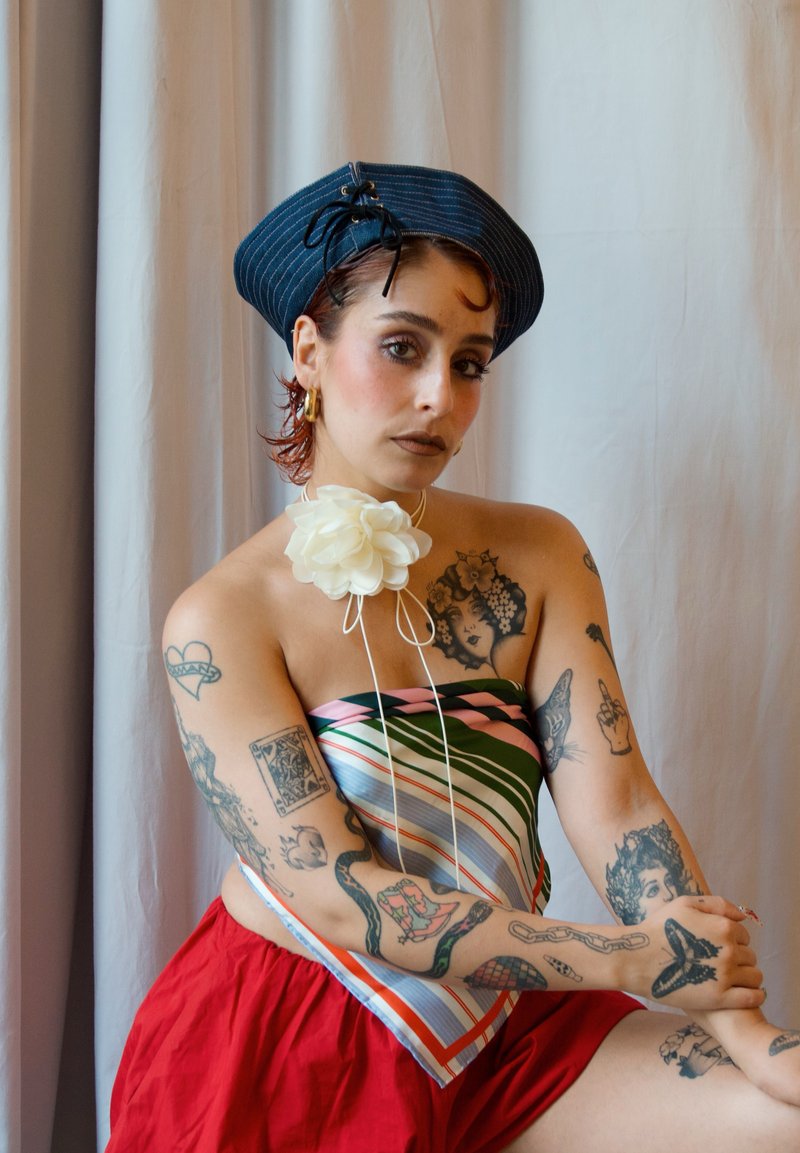 Sininen raidallinen baretti, monivärinen raidallinen hihaton toppi, punaiset hameet; valkoinen kukka-akseli kaulassa; erilaisia tatuointeja käsissä, pehmeät kankaat.