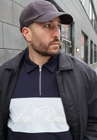 Άνδρας με σκουλαρίκια, κρίκο στη μύτη και γενειάδα φοράει γκρι καπέλο, μπλε σκούρο και λευκό πουκάμισο με φερμουάρ και μαύρο μπουφάν, στέκεται έξω από αστικό κτίριο.