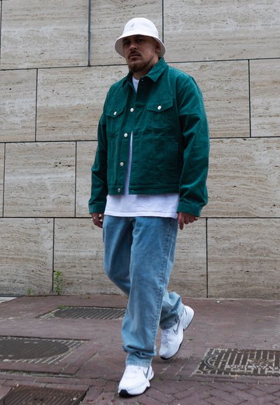 Chaqueta de mezclilla verde, camiseta blanca, jeans claros holgados y zapatillas blancas. Sombrero de cubo blanco con un logotipo. Fondo urbano pavimentado.