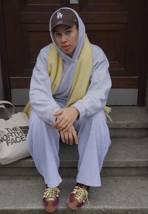 Sudadera y pantalones en gris claro, combinados con una bufanda amarilla. Gorra marrón con el logo "LA". Zapatillas multicolores. Bolsa tote al lado.