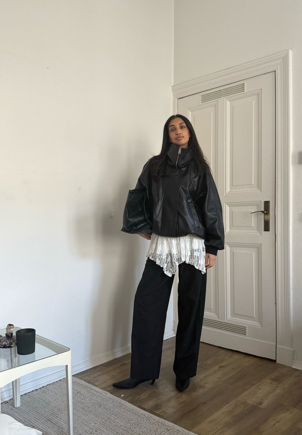 Svart skinnjakke med høy krage, lag på lag over en hvit bluse med blonder, kombinert med løstsittende svarte bukser og spisse svarte hæler, i et minimalistisk miljø.