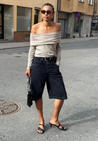 Ένα άτομο φοράει μια μπεζ μπλούζα με ανοιχτούς ώμους και μακριά μανίκια, σκούρες τζιν κοντές παντελόνες, μαύρες σαγιονάρες, και κρατάει μια μικρή μαύρη τσάντα χεριού.