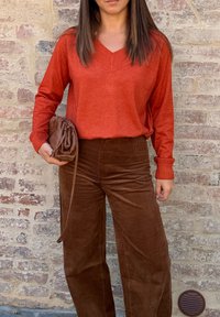Frau in einem orangefarbenen V-Ausschnitt-Pullover und braunen Cordhosen, die eine braune gewebte Clutch-Tasche hält und vor einer Backsteinmauer steht.