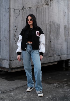 Mujer con cabello largo y oscuro que lleva una chaqueta varsity en blanco y negro, top corto negro, jeans azules, zapatillas y gafas de sol, de pie contra una pared de concreto.