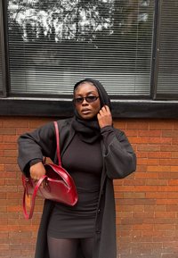 Cappotto nero lungo sopra un vestito nero, abbinato a una sciarpa nera. Tiene una borsa rossa in pelle. Sfondo con muro di mattoni grigi e persiane.