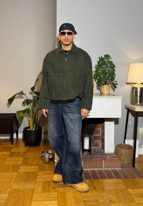 Muž so slnečnými okuliarmi, čiernou bandanou, zelenou bundou, širokými džínsami a svetlohnedými topánkami stojí v miestnosti s drevenou podlahou a izbovými rastlinami pri krbe.