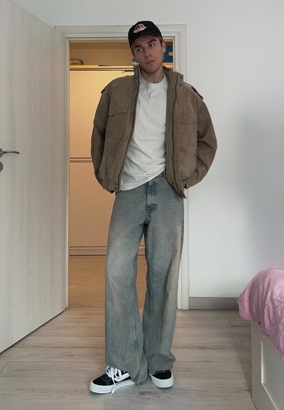 Joven con chaqueta marrón, camiseta blanca, vaqueros anchos descoloridos, gorra negra y zapatillas blancas y negras, de pie en un umbral en el interior.