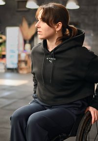 Jeune femme en fauteuil roulant portant un sweat à capuche noir Puma Hyrox, ayant un regard concentré dans une salle de sport intérieure.