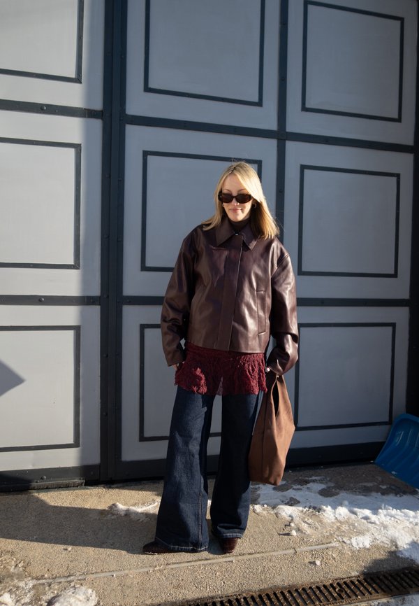 Femeie blondă purtând o geacă de piele maro închis, bluză bordo din dantelă, blugi evazați, ochelari de soare, ținând o geantă maro, stând în fața unei uși gri.