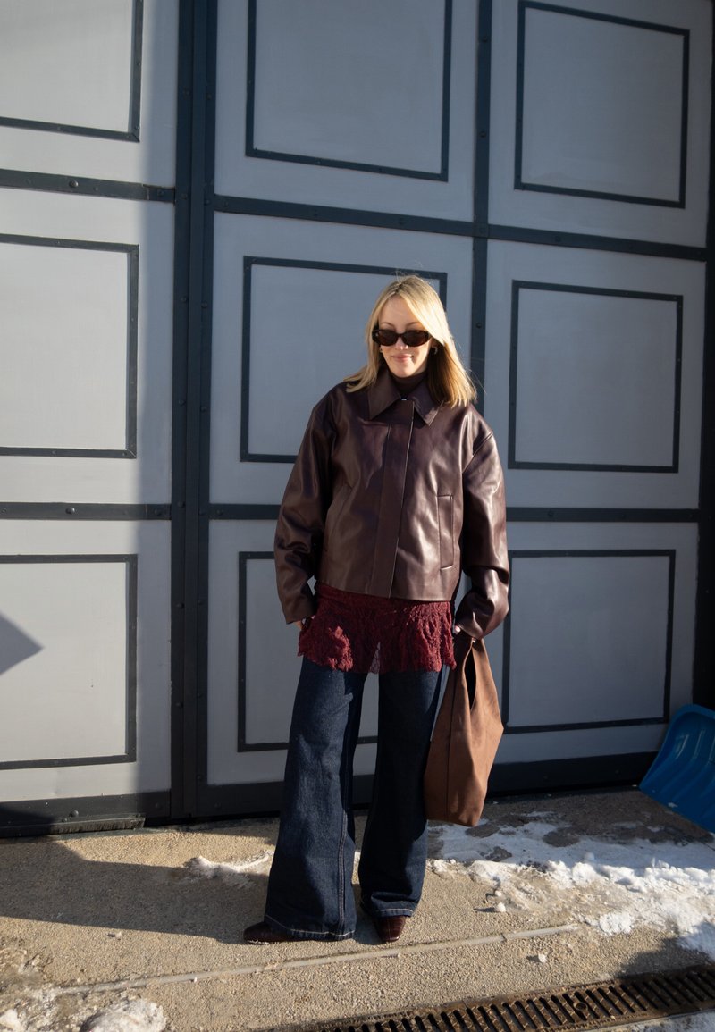 Femme blonde portant une veste en cuir marron foncé, un haut en dentelle bordeaux, un jean à jambes larges, des lunettes de soleil, tenant un sac marron, debout devant une porte grise.