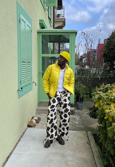 Chaqueta acolchada amarilla, camisa blanca y pantalones con estampado de vaca. De pie sobre la acera junto a un marco de puerta verde y plantas, con un cielo nublado arriba.