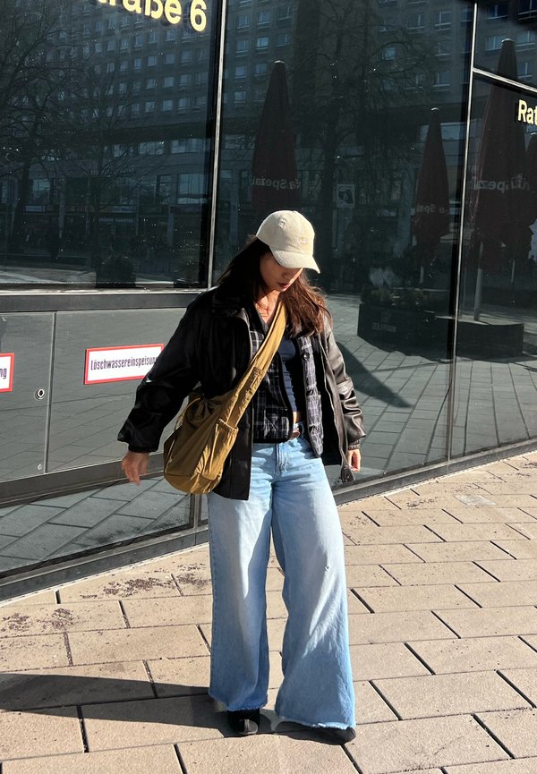 Ung kvinde iført en beige kasket, sort jakke, brede lyse jeans og en brun skuldertaske, der går på en byfortov nær reflekterende glasvægge.