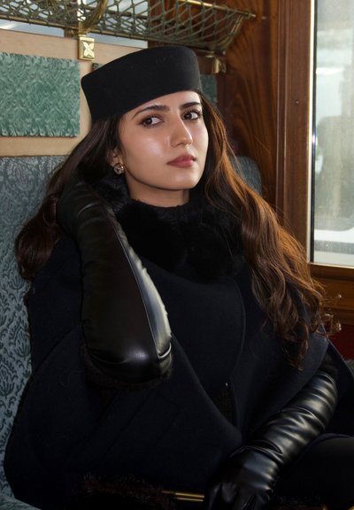 Mujer joven sentada junto a una ventana, lleva un sombrero negro, un abrigo largo negro y guantes largos de cuero negro, apoyando su mano cerca de la cabeza.