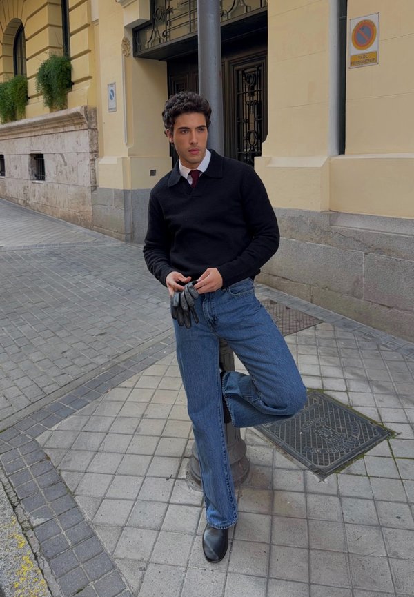 Fiatal férfi fekete pulóverben, piros nyakkendővel és kék farmerral kesztyűt tart, miközben egy városi járdán egy közvilágítási lámpának dől.