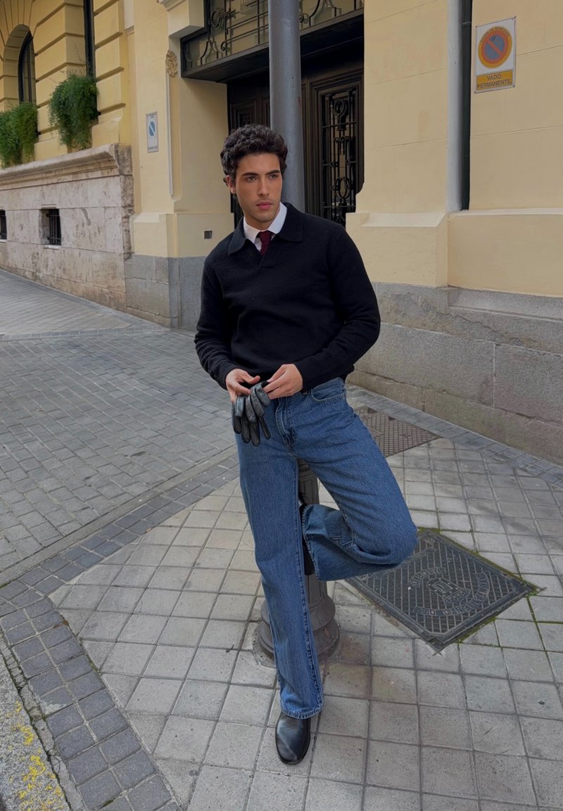 Jeune homme en pull noir, cravate rouge et jean bleu tient des gants, appuyé contre un lampadaire sur un trottoir en ville.