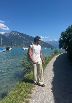 Hombre vistiendo una camiseta sin mangas blanca y pantalones anchos beige, sosteniendo una bolsa de color claro, de pie junto a un lago azul con montañas de fondo.