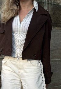 Kvinde iført en mørk cropped jakke over en hvid stribet zip-front blouse, parret med hvide bukser med ribbede sider og metalnitter.